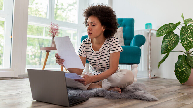 A woman sits cross-legged on a fuzzy rug on a wood floor with her laptop open in front of her. She's looking at a piece of paper she's holding up. A bright turquoise arm chair, houseplant and window are behind her. Trees and sunlight fill the window.