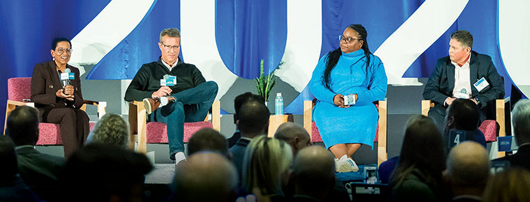 A panel of four speakers sits on stage in front of a large “2026” backdrop, speaking to an audience during a conference.