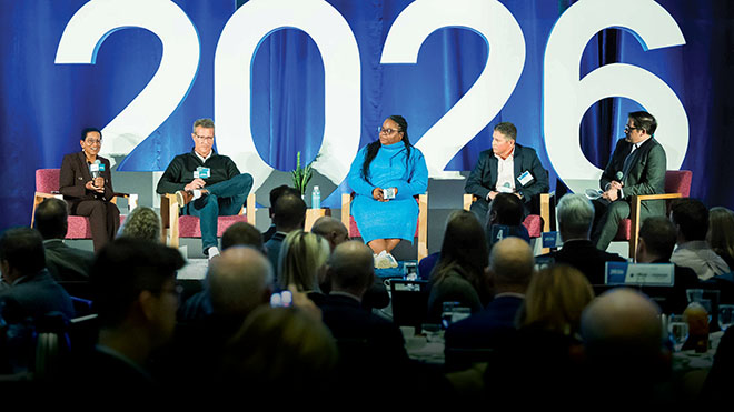 A panel of five speakers sits on stage in front of a large “2026” backdrop, speaking to an audience during a conference.