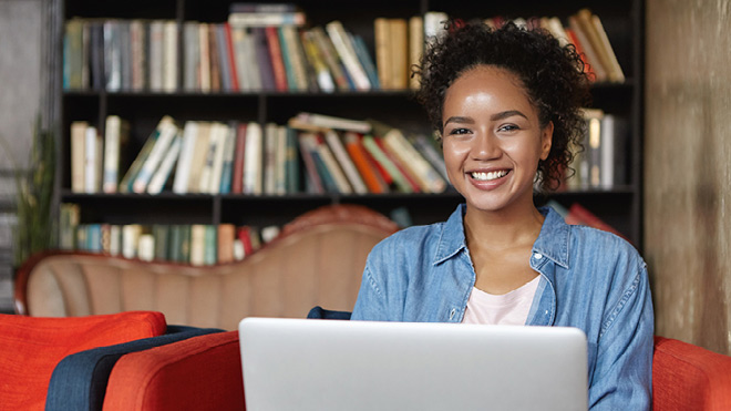 A young woman smiles at the camera over the top of her laptop screen. She is sitting on a soft orange chair. Bookshelves full of books line the wall behind her. The words BECU blog are on the left of the image.