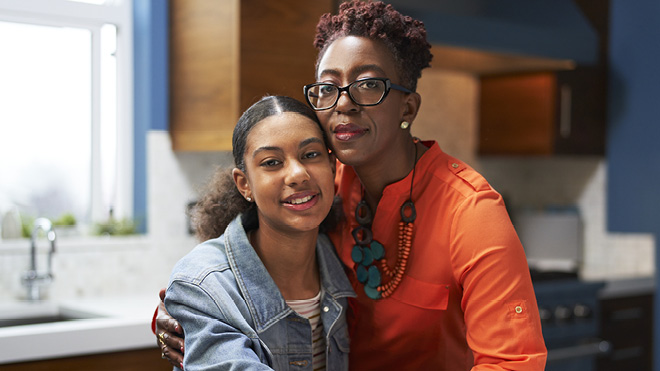 A mother and her teen daughter stand together in their kitchen facing the camera for a photo. The mom has her arm around the teen and is resting her cheek against her daughter's head.