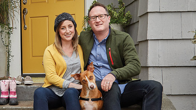 A smiling man and woman sitting in front of their home with their dog.