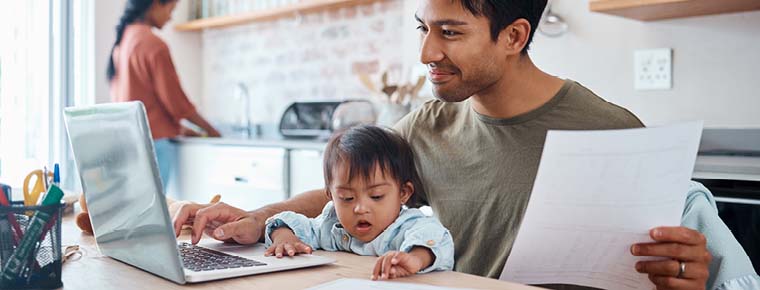 Image is of young dad looking at laptop while baby sits on his lap in the kitchen. A woman is in the background.