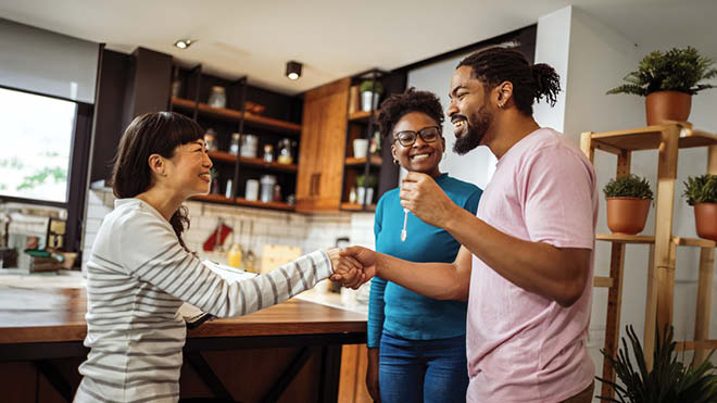 Image is of three young people who appear to be in their mid to late 20s standing facing each other in a kitchen. The person in the left side of the image is smiling and offering a handshake to the person in the right side of the image. A third smiling person is in the background.