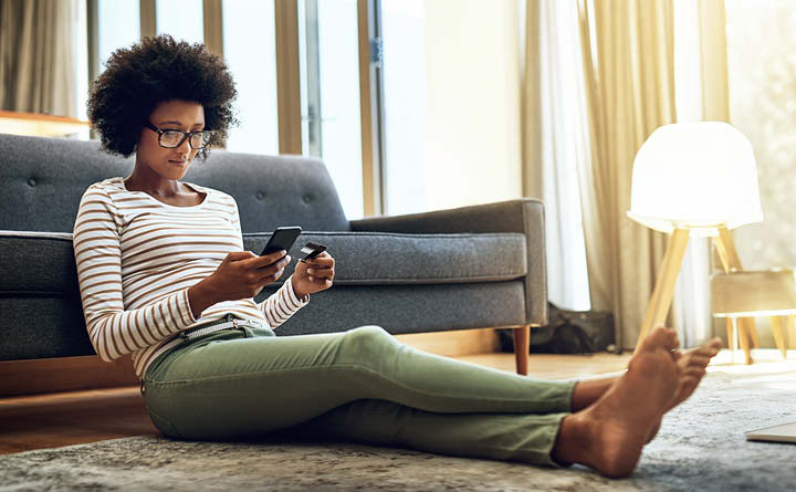 Image is of a person sitting on floor indoors. They are leaning against a couch holding a debit card and looking at a smartphone.