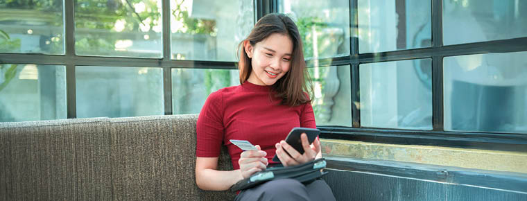 Woman sitting inside near a window with her mobile phone in one hand and her ID in her other hand.