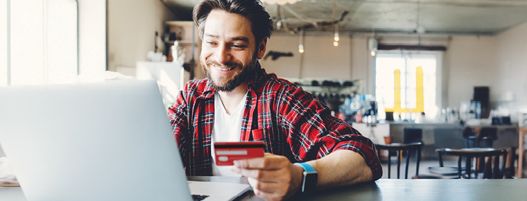 A smiling man holding his debit card and looking at his laptop.