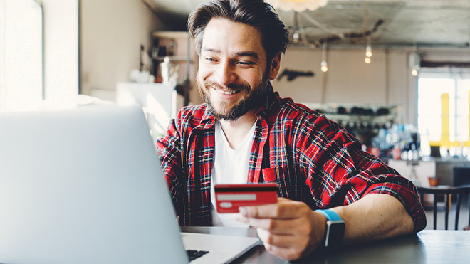 A smiling man holding his debit card and looking at his laptop.