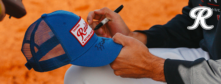 Someone signing a Tacoma Rainiers baseball cap. Tacoma Rainiers logo.