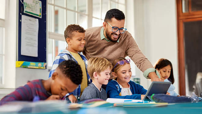 A man and a group of children are gathered around a child sitting at a table with a laptop on it, and the man is pointing at something on the laptop screen.  