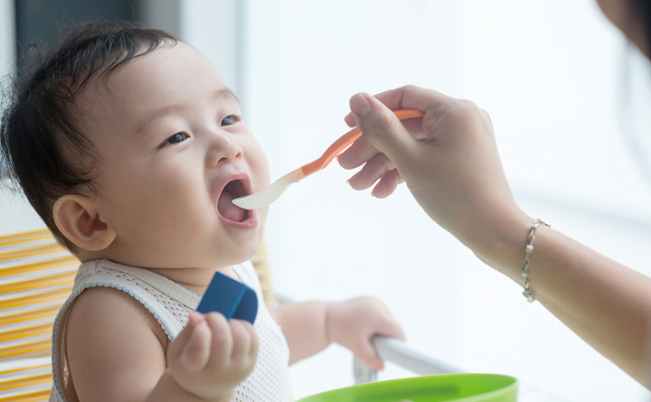 Newborn baby in a white sleeveless shirt sitting in a high chair being spoon fed food by a caregiver.