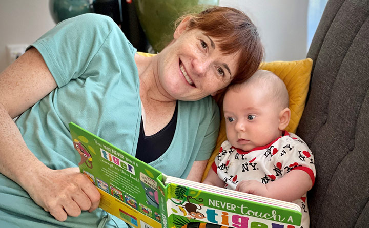 Smiling mother wearing a light green t-shirt reading the book "Never Touch a Tiger!" to a newborn baby dressed in a white, red, and black "I Love NY" onesie. The mother and newborn baby are snuggled close together on a grey couch with yellow cushions.