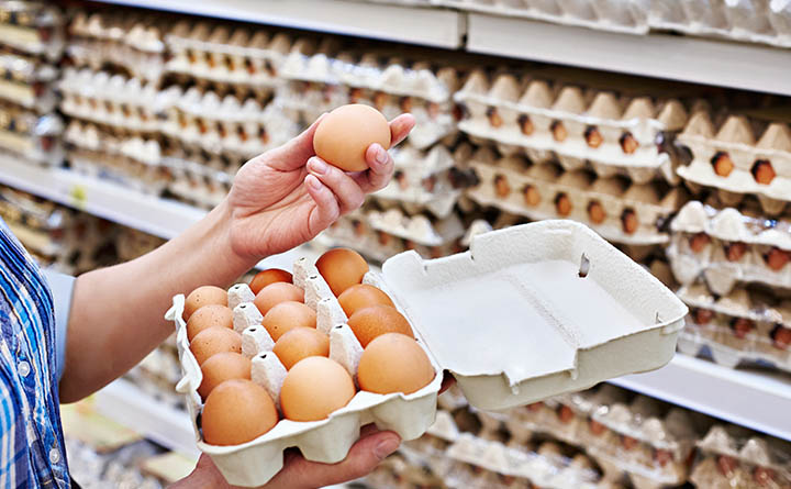 A person holds an open, cardboard egg carton of 18 brown eggs in one hand and an egg in the other. Stacks of eggs in cartons line the grocery shelves in the background.