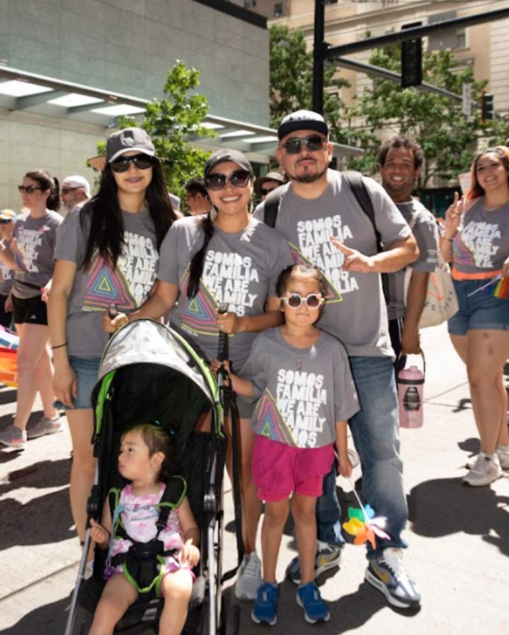Jackie Martinez-Vasquez and her family of five individuals posing for a photo on a sunny afternoon in a public setting. The entire family is wearing gray t-shirts with white text that reads: "Somos Familia. We Are Family."