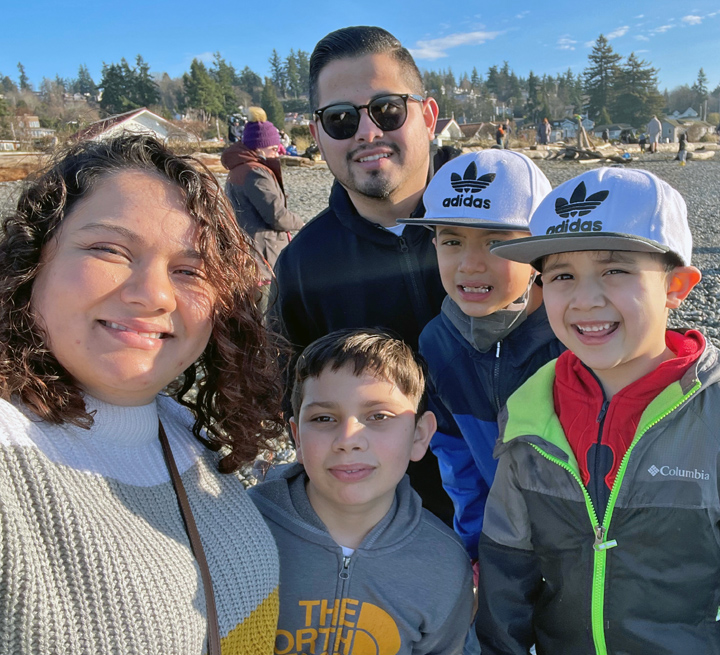 Marybeth, her husband, and their three sons, pose for a selfie at a Pacific Northwest beach, with sand, fir trees and blue sky in the background. The sun is shining and they are wearing sweaters and jackets.