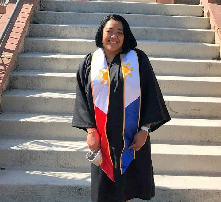Pauline Hernandez stands on steps outside a building in graduation robes and a sash representing the flag of the Philippines.