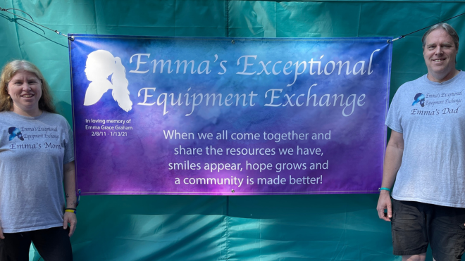 Two individuals stand next to a colorful blue and purple banner that says, "Emma's Exceptional Equipment Exchange" at the top, with a logo and slogan displaying the words "When we all come together and share the resources we have, smiles appear, hope grows and a community is made better!" A woman is on the left-hand side. She is wearing a gray shirt that says, "Emma's Exceptional Equipment Exchange" and displays the text "Emma's Mom."  On the left, a man is standing next to the banner and is also wearing the same gray shirt. His says "Emma's Dad." The two are smiling in front the banner that is hung on a teal backdrop.