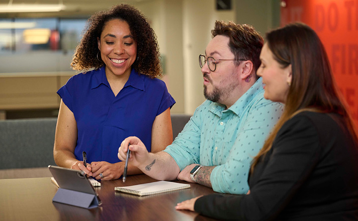 Three people are standing around a table inside a neighborhood financial center setting while looking down at a tablet. A man is in the middle. He is holding a pen and a has a notebook sitting in front of him. He is wearing a light blue long sleeve shirt and has glasses. There is a woman on the left hand side. She has curly hair. She is wearing a royal blue short sleeve shirt. She is smiling. There is also a woman on the right had side. She has long brown hair, is facing the side of the camera, and is wearing a black long sleeve shirt. In the background is a red wall and a hallway with a window.