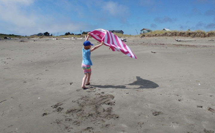 A young girl holding up a pink and white stripped towel as it flies in the air while standing on a sandy beach. She is wearing a dark blue hat, light blue shirt and rainbow-colored shorts. In the background are a few houses and blue skies. 
