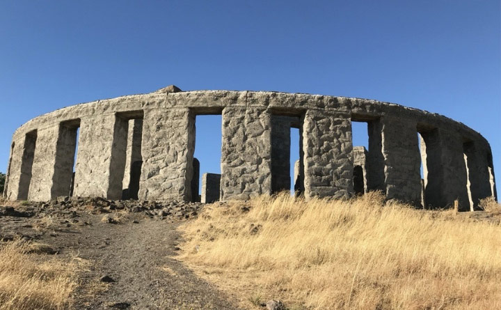A Stonehenge monument standing in a field, surrounded by dry grass and blue sky.