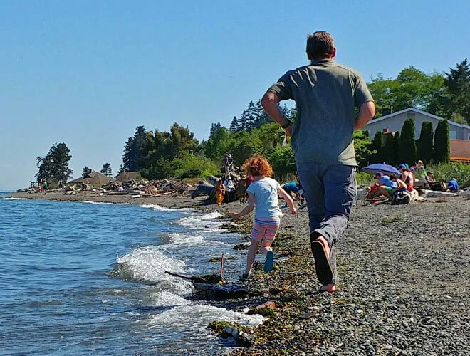 A man and a child running side by side on the beach, with the waves splashing nearby. There are others nearby sitting on the beach with an umbrella in the background. Houses and green trees are also in the background. 