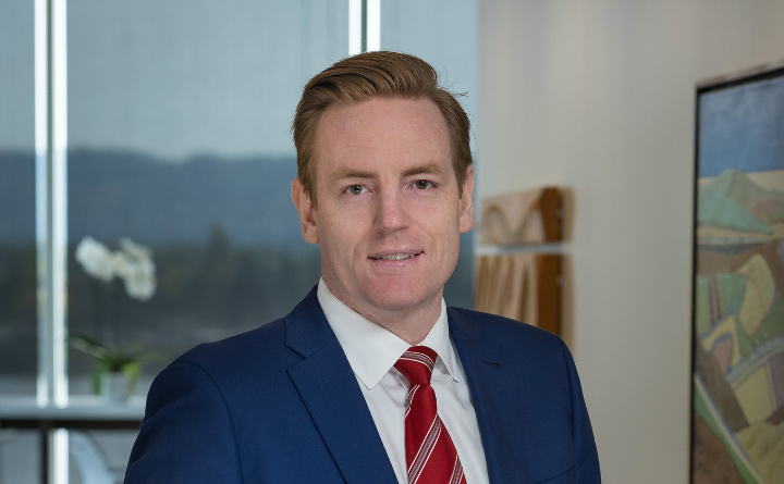 A headshot of attorney, Collin McKean. He is wearing a blue suit and red tie. He's in an office setting with a window and photography in the background.