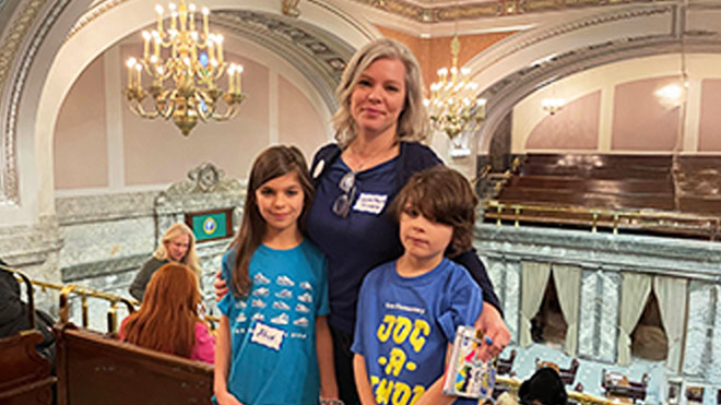 A woman and two children stand together on an interior balcony in the U.S. Capitol. Behind them are chandeliers, arched ceilings and seating. Below, the lower level is visible with curtains and additional seating.