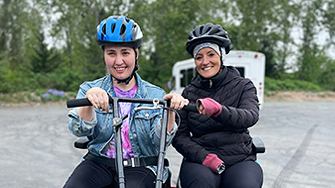 Two women sit on bikes in a parking lot, wearing helmets with their hands on the handlebars. A white bus and trees are visible in the background.