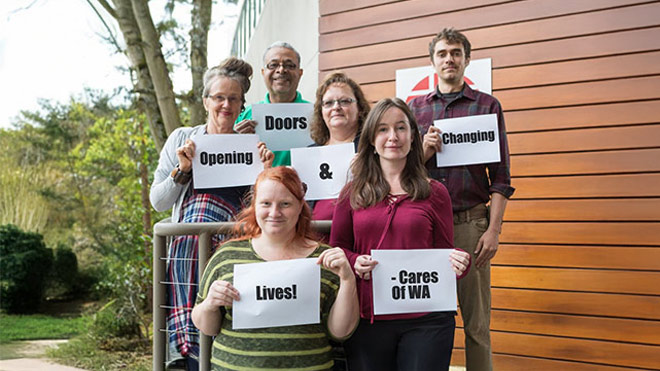 A diverse group of people stand outside next to a building, each holding a white sign. Together, the signs read: “Opening doors & changing lives! – Cares of WA.”