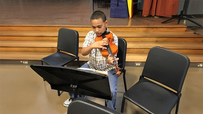 A young boy sits and plays the violin in a theater. Behind him are steps leading to a stage. He reads sheet music from a stand in front of him.