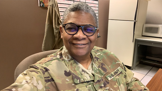 BECU employee Robin G. in U.S. Army camouflage uniform sitting in an office with a refrigerator, microwave and bulletin board in the background