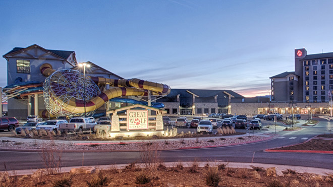 Twilight view of a resort complex featuring a hotel building, colorful water slides and a parking area. A sign reads ‘Great Wolf Lodge’ with lights shining on it.