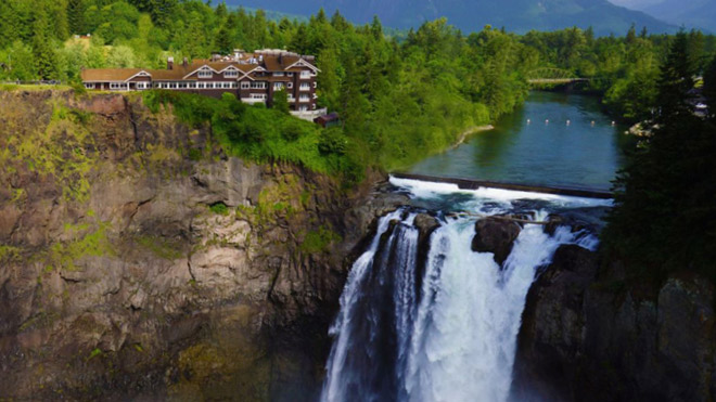 Snoqualmie Falls with the Salish Lodge perched on a rocky cliff above the waterfall. The lodge overlooks the falls, surrounded by dense green trees, with mountains visible in the background.