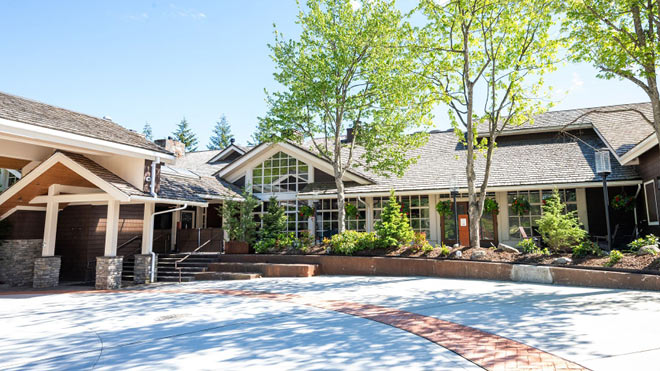 Exterior view of a lodge-style building with trees and landscaped greenery in front under a clear blue sky.