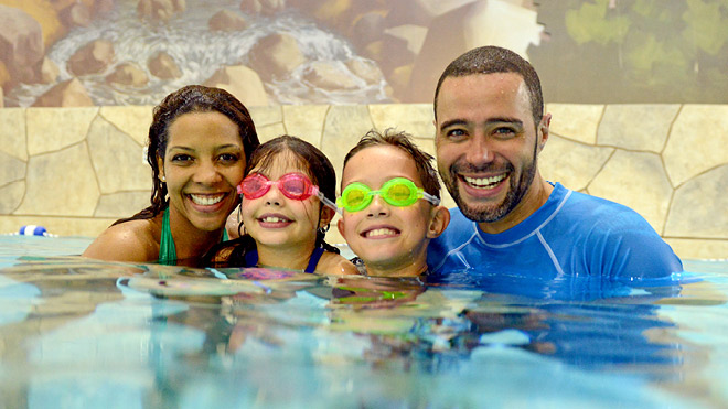 A family enjoying an indoor swimming pool with a mural of a forest scene in the background. Two kids are wearing goggles.