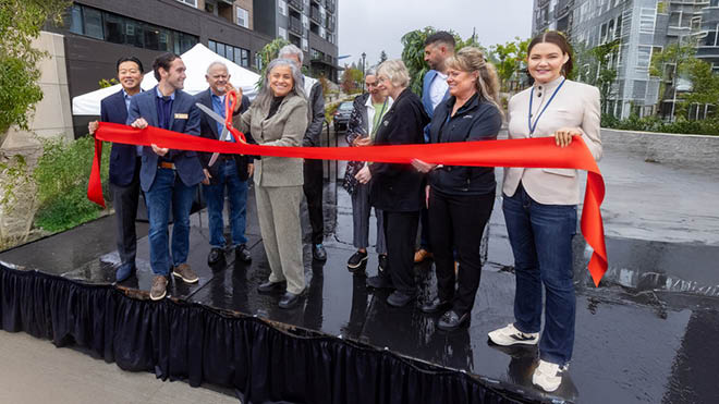 People gathered on a black outdoor stage at the Solera-Astra Apartments grand opening, holding a red ribbon and cutting it with oversized red scissors. Apartments and trees are visible in the background.