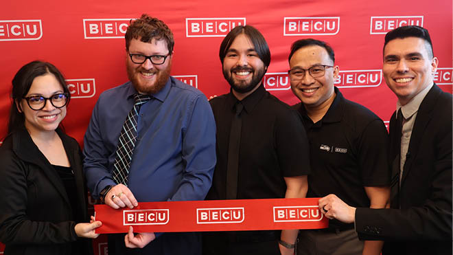 A group of five BECU employees holds a red ribbon in front of a BECU branded wall, celebrating the White Center Neighborhood Financial Centers' grand opening.
