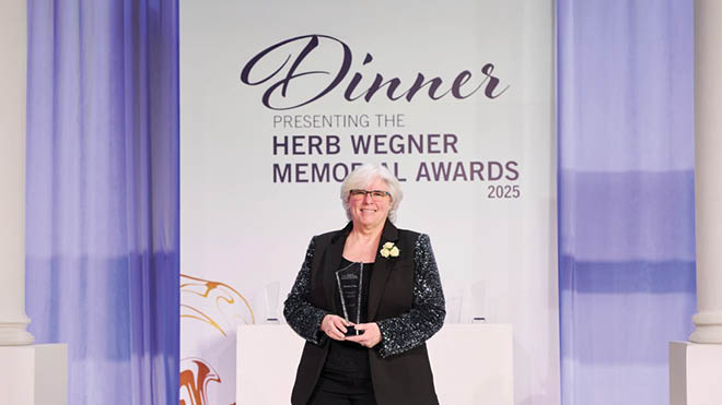 BECU employee Debbie Wege poses in front a white and purple sign that says, "Dinner Presenting Herb Wegner Memorial Awards 2025." She is holding a glass award.