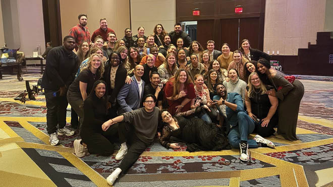 More than 30 people pose for a group photo in a hotel/conference room setting. 