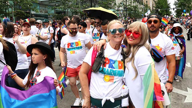 BECU employees and their family members walk down a Seattle street in the Pride parade wearing matching white shirts that say "Pride Marches On" in bright colors. Two people in the foreground stand together and smile at the camera.