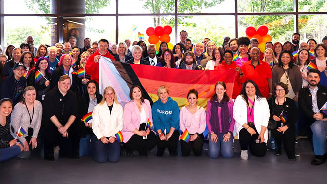 More than 60 BECU employees are gathered for a photo with a large Pride flag. Many hold small rainbow flags.