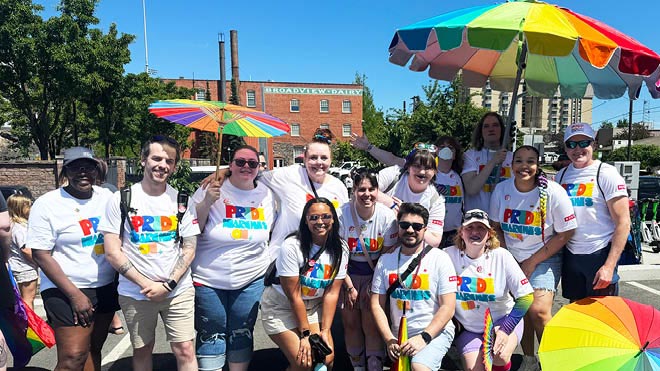 A group of 13 BECU employees wearing matching white shirts with bright lettering that says "Pride Marches On" gather for a group photo. One person is holding a small rainbow parasol. Another is holding a large rainbow umbrella.