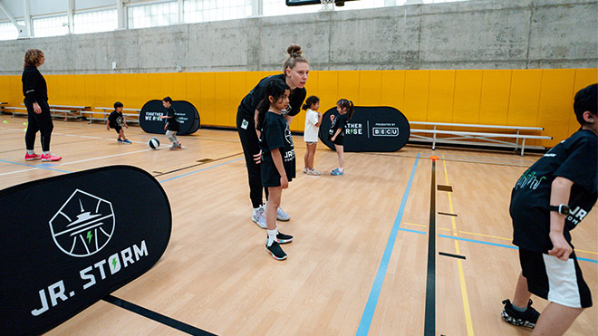 A group of children play basketball in a gym as a woman coaches alongside them. Pop-up banners on the court read 'Jr. Storm.'