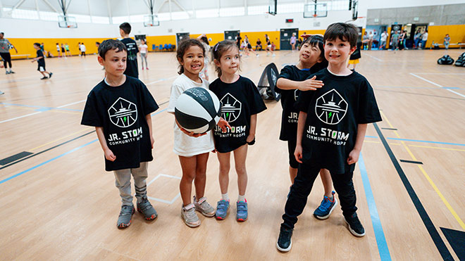 A group of children, most in black shirts, stand together in a gym, smiling and engaged in a team activity. One child holds a basketball. In the background, other kids participate in a basketball clinic.