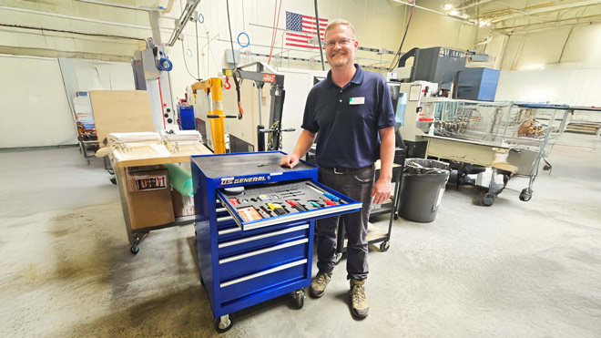 A man poses next to a large blue wheeled toolbox in a factory or machinist setting, with tools and machinery in the background.