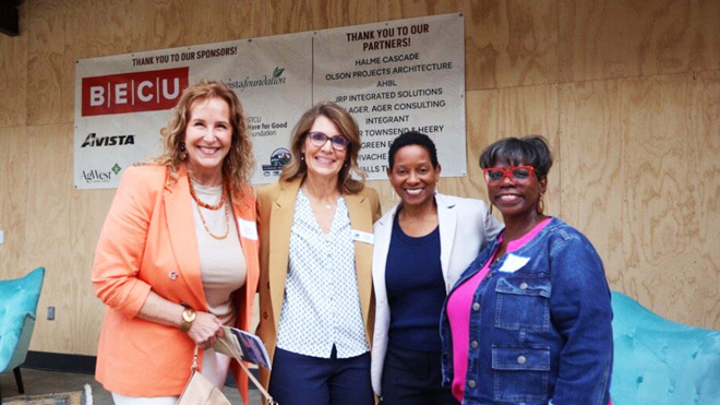 Four women stand together in front of a colorful banner, smiling and posing for the camera. The banner says, "Thank you to our sponsors" and "Thank you to our partners" and lists several organization names and logos. 