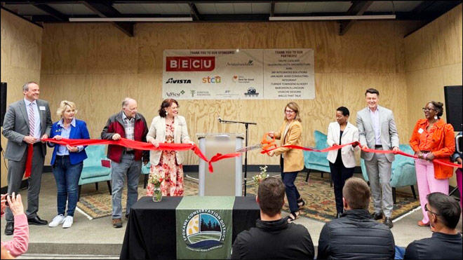 Eight people smile as a woman cuts a ribbon during a ceremonial ribbon-cutting event. Behind them, a bright banner reads 'Thank you to our sponsors,' with logos and names displayed. A podium with a microphone stands at center stage, and below the stage is a table featuring the Spokane Conservation logo. A few people in the crowd take photos and celebrate.