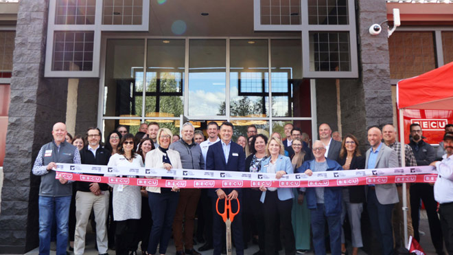 A diverse group of over 30 people pose in front of a BECU building. A man in the center holds scissors, poised to cut two ribbons. They are celebrating the opening of a new BECU Neighborhood Financial Center.