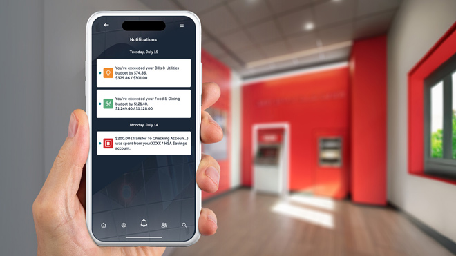 A hand holds a smartphone displaying BECU budget alerts for exceeded spending. In the background is the lobby of a BECU neighborhood financial center.
