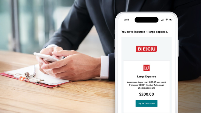A person wearing a dark business suit, sitting at a wood table, holds a smart phone in his his hands. His face is not visible. In the foreground, a phone screen shows a large-expense alert, with BECU logos. The message says: You have incurred 1 large expense. Large Expense: An amount larger than $100.00 was spent from your XXX *Member Advantage Checking account. $200.00. A teal button below the message says Log In To Account in white text.